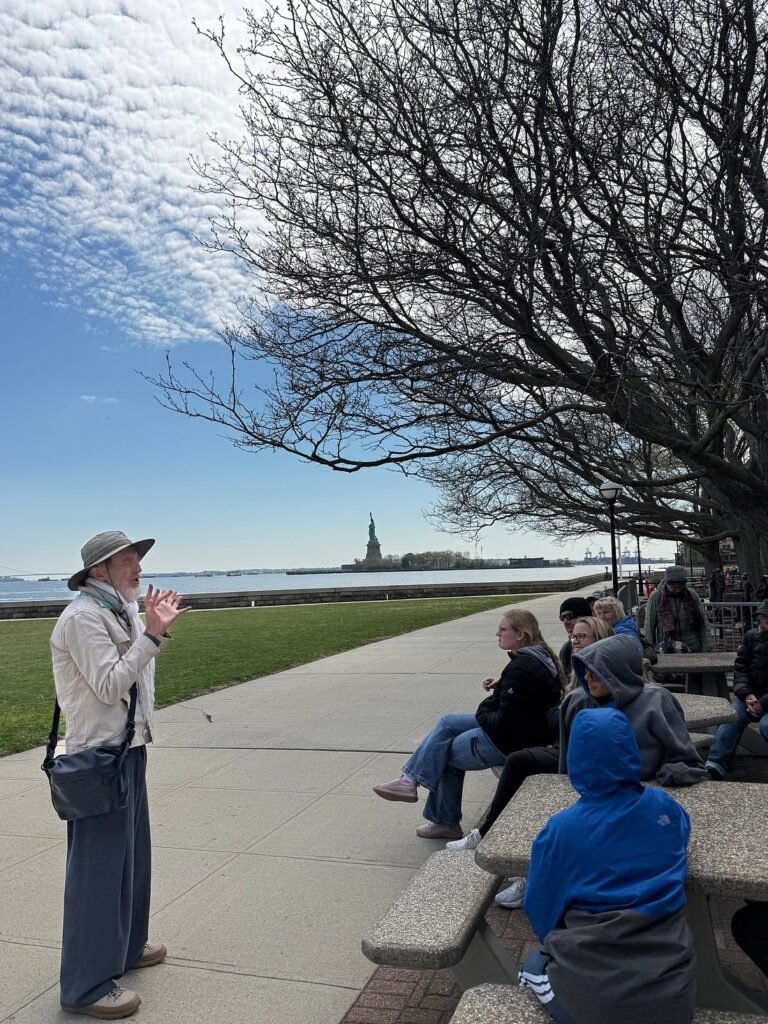 Tour guide explaining the history of the Statue of Liberty to visitors at Battery Park, with the iconic monument visible across New York Harbor, highlighting an educational sightseeing experience before boarding the Liberty Island ferry.