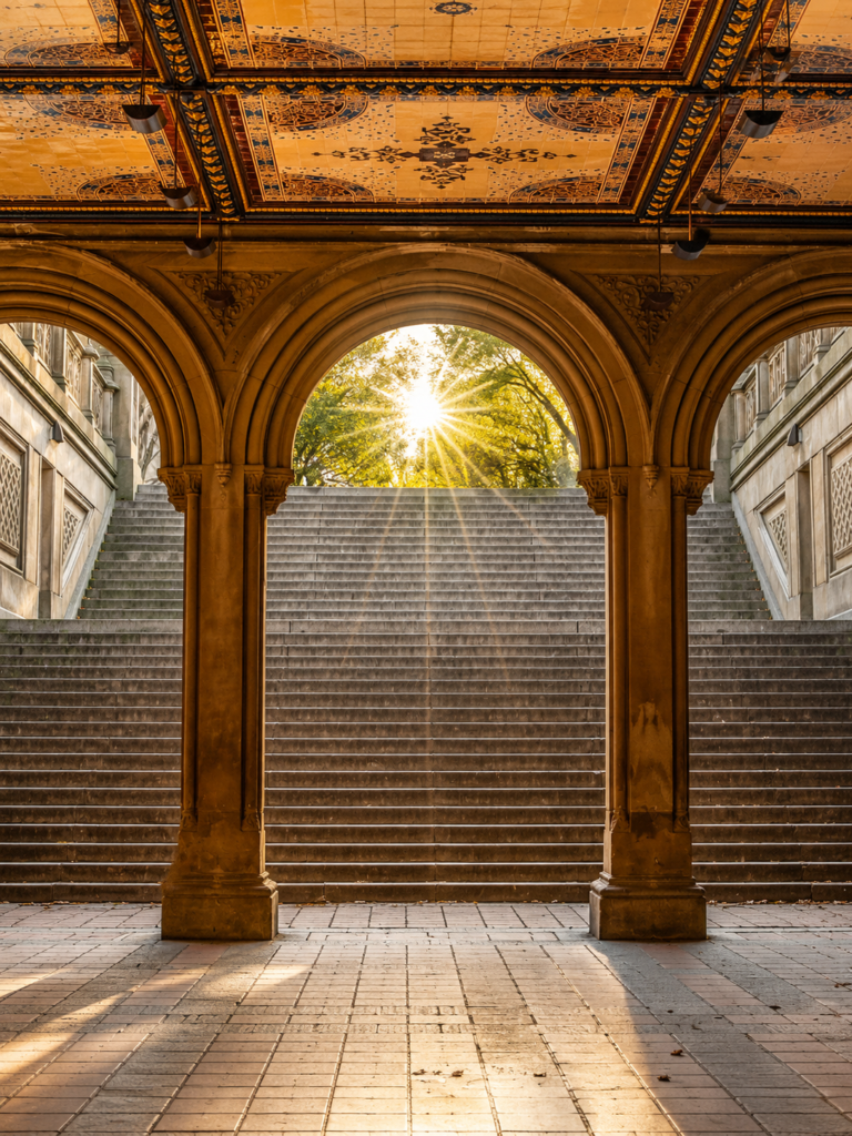 Symmetrical view beneath an ornate tiled arcade with grand stone staircases, glowing sunrise through central arch, warm golden light, intricate architectural details, historic columns, empty foreground, serene, elegant atmosphere.