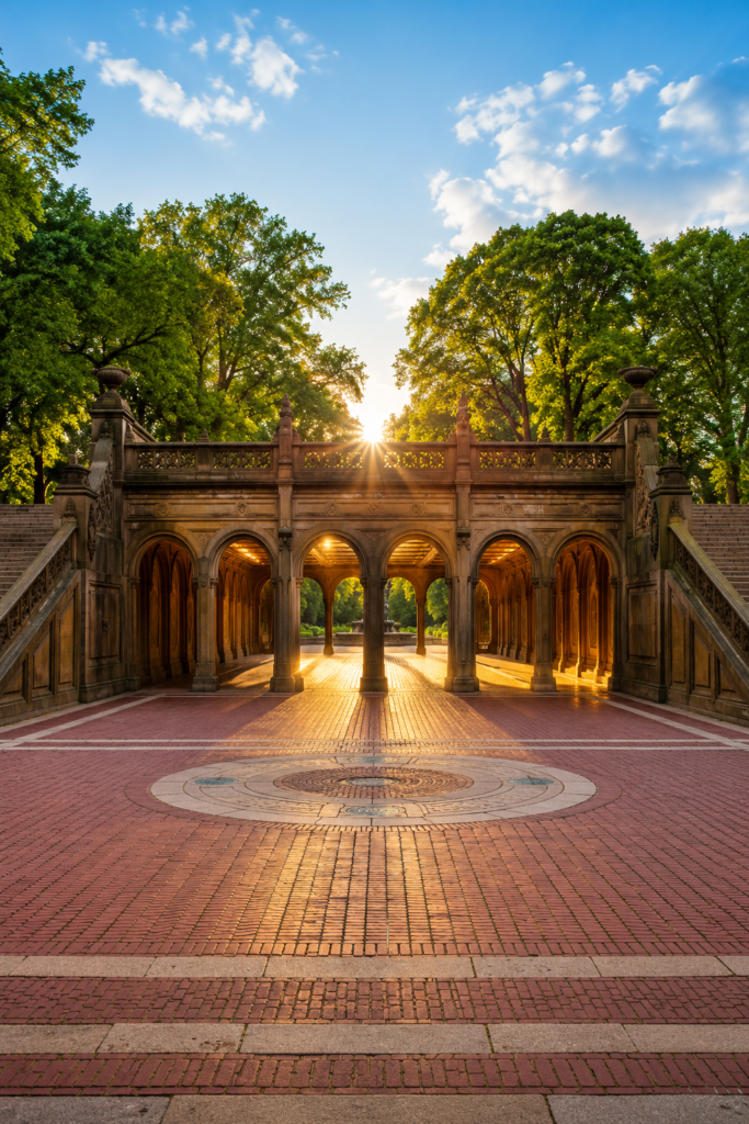 Bethesda Terrace in Central Park captured in a dramatic vertical composition, featuring elegant sandstone arches, grand staircases, intricate architectural details, glowing golden-hour sunlight, expansive brick plaza, lush green trees, and the iconic historic arcade creating a timeless, welcoming atmosphere.