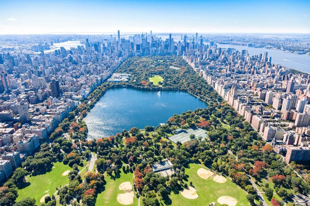 Aerial view of Central Park surrounded by the Manhattan skyline, showcasing lakes, green spaces, sports fields, and iconic New York City scenery.