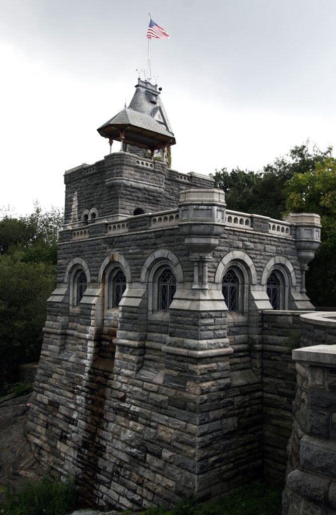 Belvedere Castle rises above rugged stone walls in Central Park, featuring historic architecture, an American flag, surrounding trees, and dramatic elevated views under overcast skies.