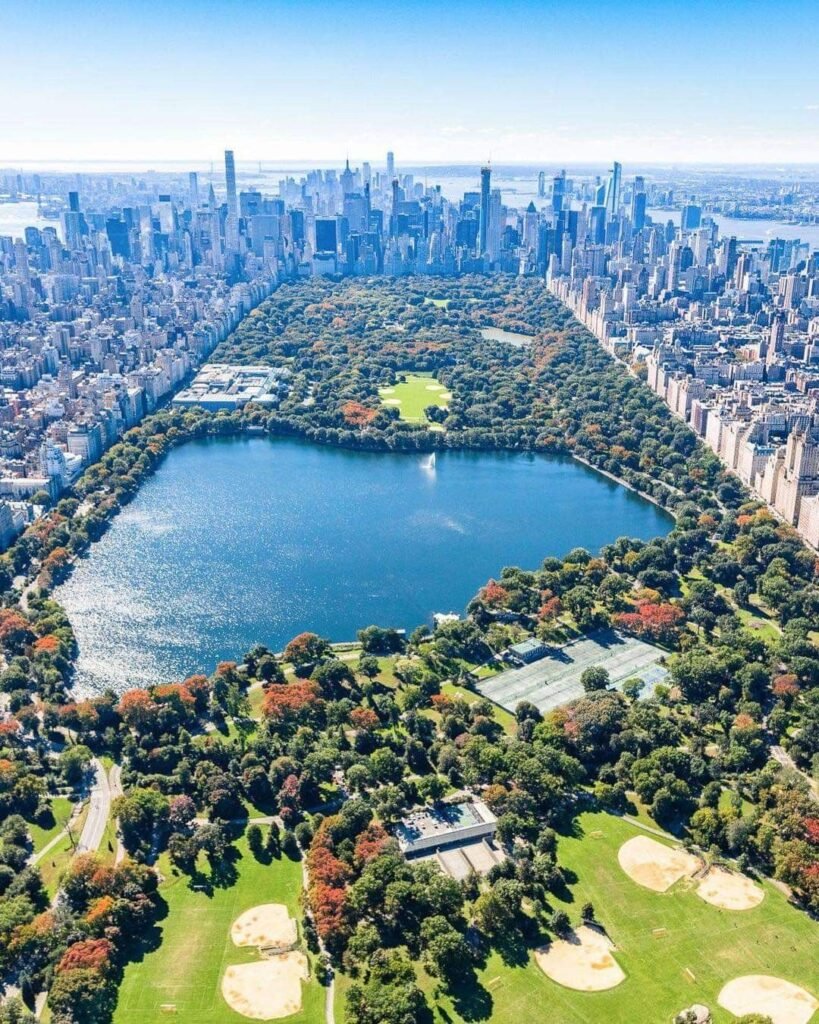 A breathtaking aerial view of the Jacqueline Kennedy Onassis Reservoir in Central Park showcases shimmering blue water, lush tree-lined landscapes, surrounding Manhattan skyscrapers, recreational fields, and the park's iconic contrast between nature and New York City's skyline.