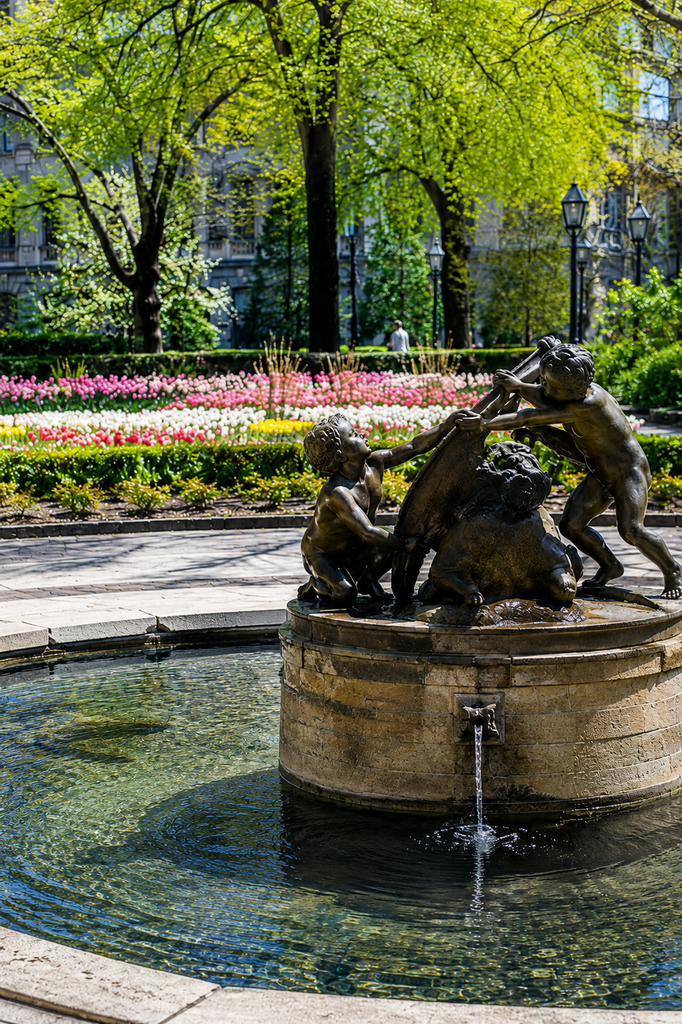 Untermyer Fountain in Central Park surrounded by colorful tulips, mature trees, and landscaped greenery, featuring a bronze sculpture above a stone fountain basin with flowing water, photographed in bright spring daylight with a classic New York park atmosphere and elegance.