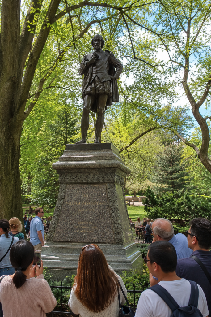 Visitors gather around the William Shakespeare statue in Central Park, surrounded by lush trees, enjoying photography, sightseeing, peaceful gardens, and sunshine.