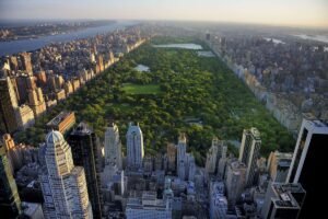 Aerial view of Central Park stretching through Manhattan, surrounded by dense city buildings, with lakes, woodlands, lawns, and streets visible under soft golden evening light across New York City skyline.